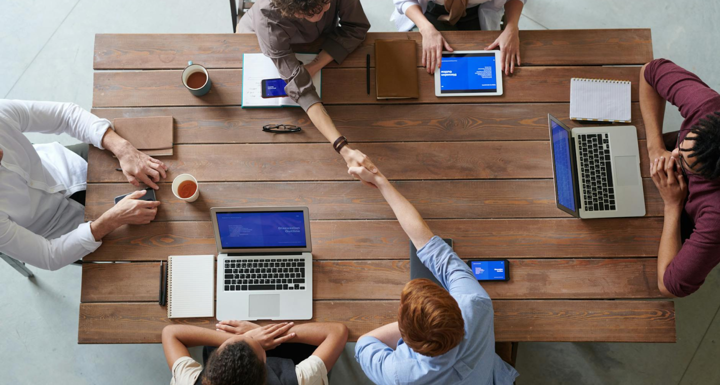 Overhead view of colleagues discussing work around a large wooden table with laptops and coffee cups