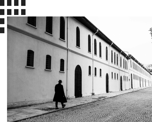 Black and white photo of a person in a trench coat walking past a long, historic building